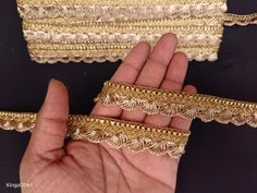 the hand is holding some gold lace on it's fingers and two pieces of fabric are