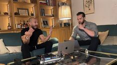 two men sitting on couches with laptops in front of them, one holding his hands up