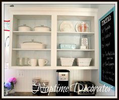 a kitchen with white cupboards filled with dishes and cups next to a chalkboard