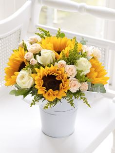 a sunflower and roses arrangement in a white bucket on a table by a window