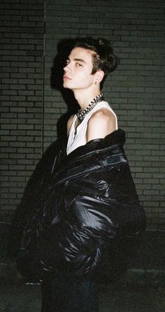 a young man in a black jacket and tie standing next to a brick wall at night