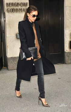 a woman in black coat and heels walking down the street with her hand on her hip