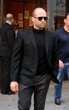 a bald man in a black suit and sunglasses walking down the street with his hand in his pocket