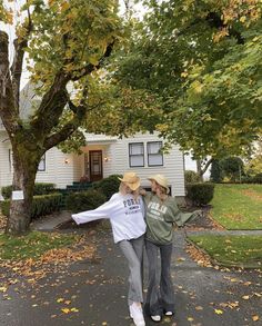 two women walking down the street in front of a white house with trees and leaves