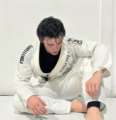 a young man sitting on the ground in white karate gear and holding his leg up