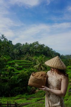 a woman wearing a straw hat holding two baskets in her hands while standing on the side of a hill