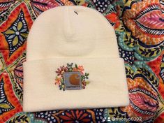 a white beanie hat with an embroidered patch on the front and side, sitting on top of a colorful blanket