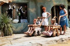 three women and two children are standing in front of a fake house with wooden toys