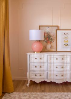 a white dresser topped with a pink lamp next to a painting and framed pictures on the wall