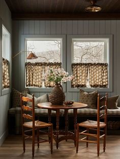 a table and chairs in a small room with two windows above the couches that have curtains on them