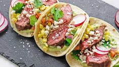 three steak tacos with corn, tomato and cilantro on a slate surface