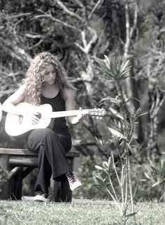 a woman sitting on a park bench playing an acoustic guitar in the grass with trees and bushes behind her