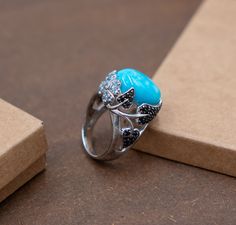 Indulge in the botanical elegance of this 10.4 gram silver ring from Gemoholic, featuring a generously sized round turquoise center stone. Flanked by intricate flower designs adorned with blue topaz stones and leaf patterns inlaid with black spinel, this ring beautifully fuses nature-inspired elements with sophisticated craftsmanship. Perfect for those who appreciate a touch of floral beauty and the deep, vibrant hues of turquoise and topaz. Turquoise Center Stone: A symbol of tranquility and wi Turquoise Flower Ring Gift, Turquoise Rings With Gemstone Accents For Gift, Turquoise Topaz Ring With Gemstone Accents As Gift, Elegant Turquoise Ring With Natural Stones For Anniversary, Elegant Turquoise Rings With Natural Stones, Silver Turquoise Ring, Leaf Patterns, Turquoise Ring Silver, Turquoise Flowers