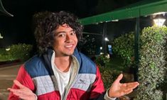 a man with curly hair standing in front of a building at night, holding his hands out