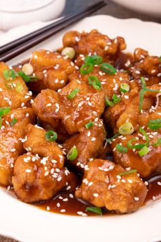 a white plate topped with chicken covered in sauce and sesame seeds next to chopsticks