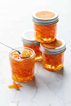 four jars filled with orange peels on top of a white marble counter next to a spoon