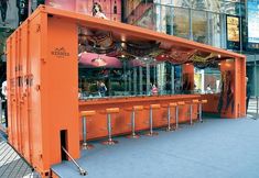 an orange bar on the side of a street with people walking by it and buildings in the background