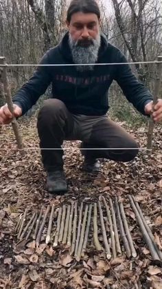 a man squatting down on the ground with sticks in front of him and trees behind him