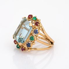 Stylish vintage aquamarine & emerald cocktail ring (circa 1950s to 1960s) crafted in 14 karat yellow gold.   Emerald cut aquamarine measures 16.5mm x 14mm (estimated at 16.50 carats), accented with an estimated 1.80 carats of cabochon cut emeralds, blue sapphires and rubies. The aquamarine is in very good condition and free of cracks or crisps.  The sky blue aquamarine is securely set in a four pronged mount, bordered with a sweets pop setting of rubies, emeralds and blue sapphires. A striking a Formal Multi-stone Aquamarine Rings, Cocktail Jewellery, Emerald Cocktail Ring, Emerald Cocktail, Gemstone Rings Vintage, Goddess Jewelry, Athena Goddess, Cocktail Jewelry, Gold Cocktail