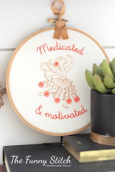 a cross - stitch hoop hanging on the wall with a plant and books beside it