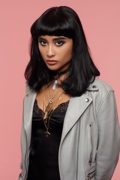 a woman with black hair wearing a leather jacket and necklaces, standing in front of a pink background