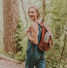 a woman in overalls with a backpack on her back walking through the woods and smiling