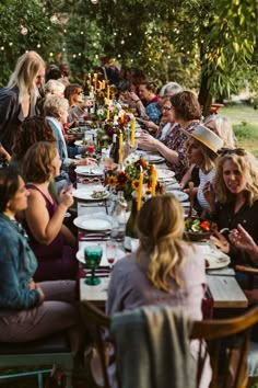 a group of people sitting at a long table eating food and drinking wine in the park