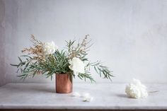 white flowers are in a copper vase on a marble countertop next to an artificial flower arrangement