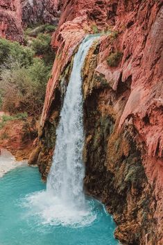 a large waterfall flowing into a river surrounded by red rocks and greenery in the desert