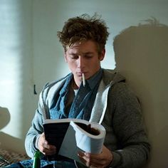 a man sitting on a bed reading a book and drinking from a paper cup in his mouth