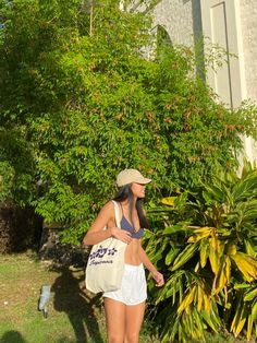 a woman in white shorts and a tan hat is holding a bag near some bushes