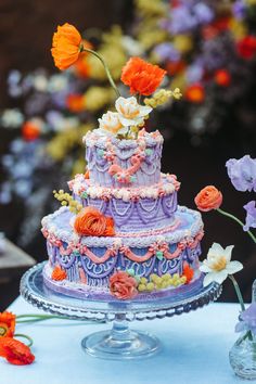 a three tiered cake decorated with flowers on a glass platter next to an orange and purple flower arrangement