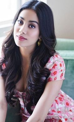 a woman with long dark hair sitting on a couch wearing a pink dress and gold earrings