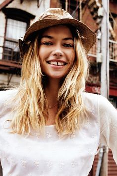 a woman with long blonde hair wearing a hat and white shirt standing in front of a building