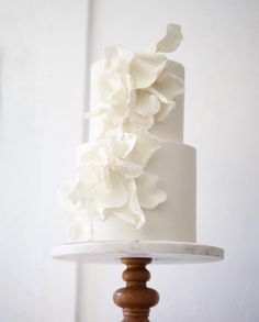 a white wedding cake with flowers on top is sitting on a wooden stand in front of a white wall