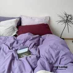 an unmade bed with purple sheets, pillows and a plant on the wall behind it