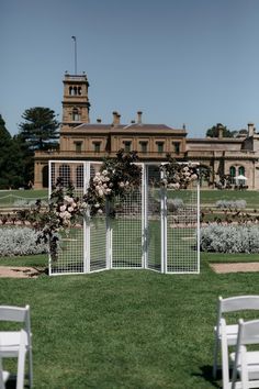 an outdoor ceremony setup with white chairs in front of a large building and flowers on the lawn