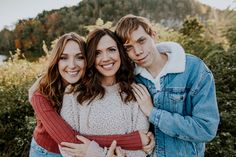 two girls and a boy are hugging each other in front of some bushes with mountains in the background