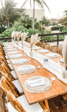 a long table set with place settings and white napkins on it, along with palm trees in the background