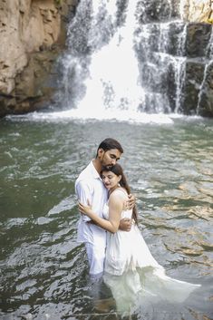 a man and woman standing in the water next to a waterfall with their arms around each other