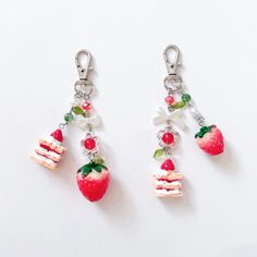 two strawberry charms are hanging from silver hooks on a white surface, one has a piece of cake and the other is a strawberry with green leaves