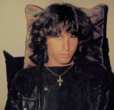 a man with long hair sitting in a chair wearing a black leather jacket and necklace