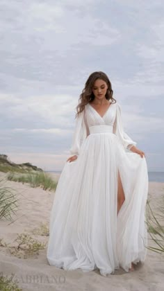 a woman standing on top of a sandy beach wearing a white dress and long sleeves