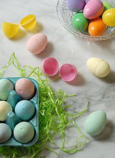 several different colored eggs in a plastic container on a table next to some grass and small bowls