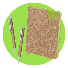 two pens sitting next to a cork covered notebook and pen holder on a green circle