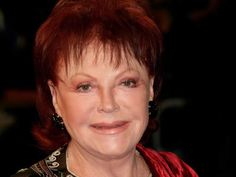 an older woman with red hair and green eyes smiles at the camera while wearing black earrings