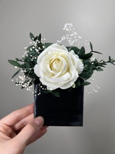 a hand holding a white rose and baby's breath in a black square vase