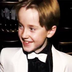 a young man wearing a white suit and black bow tie smiles at the camera while sitting in a chair