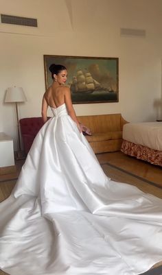 a woman in a white wedding dress is standing near a bed with a painting on the wall behind her