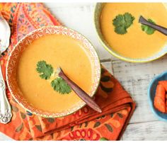 two bowls filled with carrot soup on top of a colorful napkin next to spoons